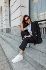 Wall Mural - Fashionable beautiful young woman business model with vintage sunglasses in a fashion black suit with a blazer and sneakers sits on the steps near a vintage building in the city