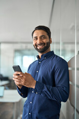 Wall Mural - Smiling Indian businessman using mobile cell phone working standing in office. Happy business man making bank transaction payment on smartphone working on cellphone at work. Vertical portrait.