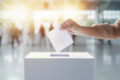 Close up hand of woman putting letter in white ballot box at modern hall in background of blurred people. Voting concept of politics and elections.