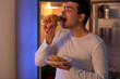 Hungry young man eating croissant near open fridge in kitchen at night