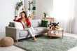 Beautiful young Asian woman with glass of wine and snacks sitting on sofa in living room