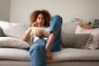 © Daniel - Young multiracial woman watching movie at home eating popcorn, sitting on the couch with surprise expression.