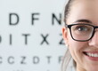© H_Ko - Focus on beautiful woman wearing glasses in black stylish frame. Cheerful ophthalmologist looking at camera with happiness. Letters table for eyesight check on background. Vision test concept