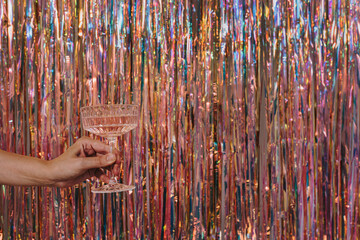 Persons hand holding glass of champagne wine over sparkling tinsel curtain. Christmas, New Year holidays party event celebration background. Foil fringe shimmer curtain