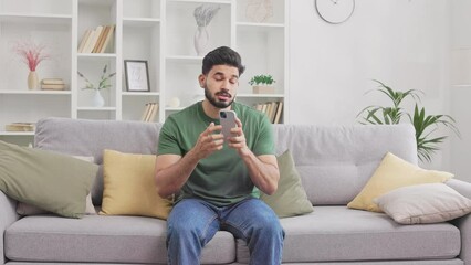 Bearded indian man feeling annoyed while having problem with mobile application at home. Casually dressed hindu male sitting on grey sofa and and getting angry.