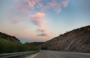 Driving on a mountain road at sunset, motion blur effect. Beautiful landscape of highway with pink clouds on the sky