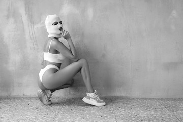 Wall Mural - Beautiful sexy woman in underwear. Model wearing bandit balaclava mask. Hot seductive female in nice lingerie posing near grey wall in studio. Crime and violence. Sits on the floor. Black and white