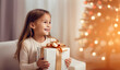 © kanina - Little girl holding a gift with a golden bow, smiling, in the background of a Christmas tree with lights, generative ai