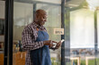 © Wasana - A healthy retired black senior man stands proudly at his new personal business. Hold a tablet to check and take orders. Startup business owner. SME entrepreneurs.