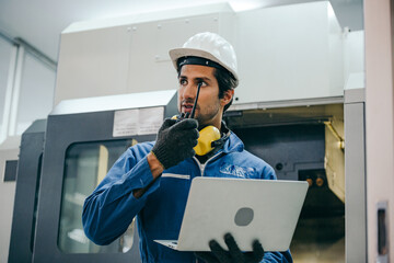 Wall Mural - Male industrial engineer or technician in safety hard helmet and uniform using laptop checking on machine and radio phone calling team worker, heavy technology invention industry manufacturing factory