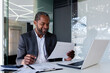 © Liubomir - A young African American male businessman is working in the office on a laptop and holding documents in his hands.