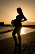 pregnant woman on the beach with backlit sunset and a hat
