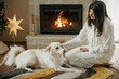 Beautiful woman in stylish pajamas relaxing with cute white dog at cozy fireplace, enjoying calm christmas morning in festive decorated living room. Merry Christmas! Winter holidays with pet