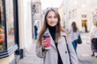 © okrasiuk - Young smiling woman in trench coat enjoying hot coffee drink in reusable cup while walking on the city streets decorated for the autumn holidays. Happy fall cozy mood, seasonal fashion style trend.