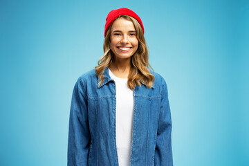 Wall Mural - Portrait of beautiful smiling woman wearing red hipster hat and stylish jeans jacket isolated on blue background. Female tourist with wavy silky hair looking at camera. Travel concept