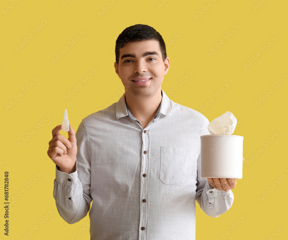 Young man with nasal drops and tissue box on yellow background