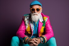 Portrait Of A Stylish Senior Hipster Man With Long White Beard And Mustache Wearing Bright Colorful Clothes And Sunglasses Sitting On A Purple Background