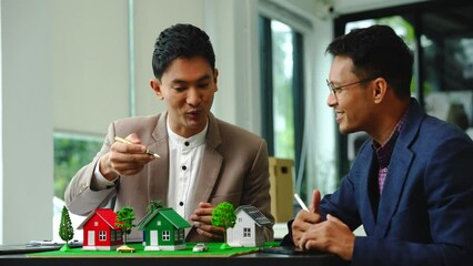 Wall Mural - Two professionals smiling, discussing over model houses on a table, possibly planning or reviewing a housing project on large land. Asian people, only businessman, Middle-age