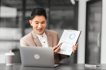 Asian professional male business person in beige suit and white shirt is working on laptop and holding pen, looking focused.