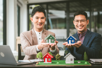 Wall Mural - Two professionals smiling, discussing over model houses on a table, possibly planning or reviewing a housing project on large land. Asian people, only businessman, Middle-age