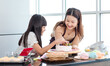 © Bangkok Click Studio - Asian beautiful female baker pastry chef mother wears apron standing smiling helping teaching little pretty girl daughter decorating cake with mixed berry fruit making homemade bakery in home kitchen