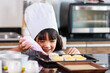 Closeup shot of Asian female little girl baker pastry bakery chef daughter wears white tall hat standing smiling using colorful whipped cream decorating preparing homemade cupcakes in home kitchen