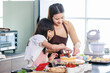 © Bangkok Click Studio - Asian beautiful female baker pastry chef mother wears apron standing smiling helping teaching little pretty girl daughter decorating cake with mixed berry fruit making homemade bakery in home kitchen
