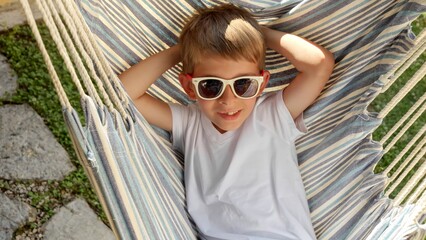 Wall Mural - At peace in a garden a boy in sunglasses swings and lounges in a hammock, expressing the sensations of summertime, the innocence of childhood, calmness, and the magic of summer holidays