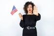 Young beautiful woman with curly hair wearing black dress and holding and American USA flag and making stop gesture over white studio background. 