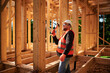 Carpenter constructing wooden frame house. Bearded man with glasses is hammering nails into the structure, wearing protective helmet and construction vest. Concept of modern ecological construction.
