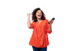 young curly brunette woman dressed in orange blouse rejoices holding phone