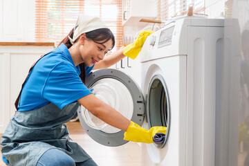 Professional Asian woman cleaning service wearing yellow rubber gloves, using a rag to wipe the washing machine near the kitchen counter at home. Housekeeping cleanup, Woman cleaner concept.