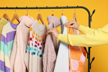 Wall Mural - Woman taking stylish sweater from clothes rack against orange background, closeup