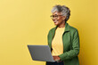 Smiling elderly African American IT woman 50s years old wears jacket white t-shirt hold closed laptop pc computer isolated on plain yellow background studio portrait. People lifestyle concept