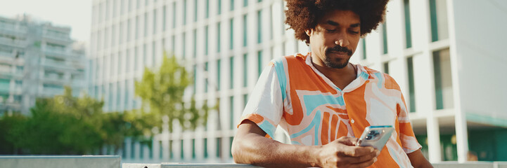 Closeup young African American man wearing shirt writes in social networks on mobile phone on urban street background. Camera moving forwards approaching to the person.