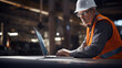 Mature construction worker or engineer is intently using a laptop on-site, wearing a safety helmet and reflective vest, with construction activity in the background.