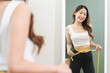 Loss weight slim, smile asian young woman measuring her waist size with tape measure, wear in fit sport, looking reflect in mirror at home. Sport strong person in gym fitness for wellbeing healthcare.