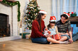 Asian young father and mother push forward daughter open gift box that parent give to her while sitting on a floor near fireplace and pine tree decoration in Christmas December festive event at home