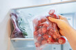 Female hand holds plastic bag with frozen strawberries against background of an open freezer.