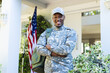 Happy african american male soldier with arms crossed in cap outside home with usa flag, copy space