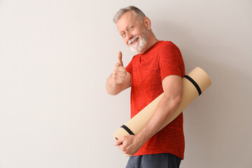 Wall Mural - Sporty mature man with fitness mat showing thumb-up on light background