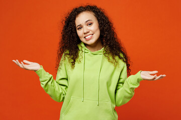 Wall Mural - Young sad doubtful woman of African American ethnicity she wear green hoody casual clothes shrugging shoulders looking puzzled spread hands isolated on plain red orange background. Lifestyle concept.