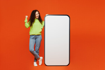 Wall Mural - Full body young woman of African American ethnicity wear green hoody casual clothes big huge blank screen mobile cell phone smartphone with area do winner gesture isolated on plain orange background.