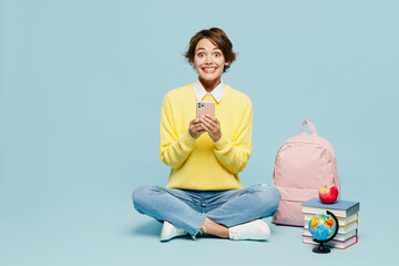 Wall Mural - Full body young fun woman student wear casual clothes sweater sit near books backpack bag hold in hand use mobile cell phone isolated on plain blue background. High school university college concept.