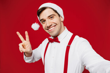 Wall Mural - Close up merry young man wear white shirt Santa hat posing doing selfie shot pov on mobile cell phone show v-sign isolated on plain red background Happy New Year Christmas celebration holiday concept