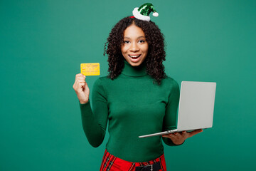 Merry fun little IT kid teen girl wear hat casual clothes posing using laptop pc computer credit bank card shopping online isolated on plain green background. Happy New Year Christmas holiday concept.