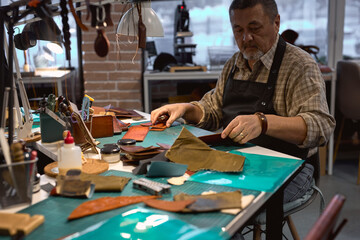 talented old serious man wearing apron and checked shirt using an awl makes holes for hand-stitching a leather product. closeup portrait.