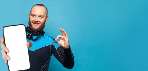 Diving guy showing okay gesture and smartphone with empty white screen display for new Application on blue studio wall background. Mockup banner