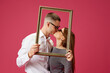 © master1305 - Portrait of happy young couple, man and woman, posing with picture frame, kissing against pink studio background. Concept of love, relationship, Valentine's Day, emotions, lifestyle