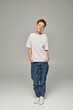 full length of happy redhead queer person in white t-shirt and jeans with hands in pockets on grey